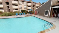 This amenity space features two distinct areas designed to support an active lifestyle: an outdoor swimming pool and an indoor fitness center. The pool area is generously sized with a curvilinear shape and clear blue water. A spacious pool deck surrounds the water, furnished with blue lounge chairs for relaxation. A shaded seating area with a blue umbrella and table provides a comfortable retreat. Lush landscaping lines the perimeter, and the multi-story residential building backdrop creates an enclosed, community-oriented atmosphere.

The fitness center is a well-lit, single-floor space distinguished by bold orange and blue accent stripes along the upper walls, lending the room an energetic character. Large windows along one wall overlook the pool area, flooding the interior with natural light. The space is carpeted with a patterned tile-style flooring and equipped with an array of cardio machines, including multiple treadmills and an elliptical trainer. Strength-training options include a multi-function cable machine, stationary bikes, a rowing machine, and a full dumbbell rack backed by a large wall mirror. A ceiling fan and recessed lighting ensure comfort throughout workouts.