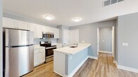 This single-floor residence offers a clean, modern aesthetic highlighted by soft gray walls, white trim, and wood-look luxury vinyl plank flooring throughout the main living areas. The kitchen is a standout feature, outfitted with crisp white shaker-style cabinetry, stainless steel appliances including a refrigerator, electric range, and over-the-range microwave, and a peninsula with a white countertop and undermount sink. The peninsula provides a natural transition into the open living area, which is generously sized and filled with natural light through sliding glass doors fitted with vertical blinds. The layout flows intuitively, with clear sightlines connecting the kitchen and living spaces. The bedroom features plush carpeting and a large window overlooking greenery, offering a quiet, comfortable retreat. The full bathroom includes a white vanity with shaker-style cabinetry, wood-look flooring, and a full tub and shower combination with built-in shelving. Throughout the space, flush-mount lighting fixtures provide consistent illumination, and ceiling vents indicate a central HVAC system. The overall design is modern, functional, and move-in ready.