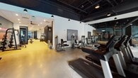 Located at 2401–2439 Old Brick Rd in Glen Allen, Virginia, this contemporary fitness center offers a well-appointed, single-floor layout designed to support a full range of workout styles. The space is divided into distinct training zones, each thoughtfully equipped and styled to enhance the exercise experience.

The cardio area features a row of treadmills and elliptical machines positioned near large windows that invite abundant natural light and outdoor views of the surrounding greenery. Deep teal accent walls and an exposed black ceiling with industrial pendant lighting give this zone a bold, modern aesthetic.

Adjacent to the cardio section, a spacious strength training floor hosts a comprehensive selection of Life Fitness selectorized machines and cable systems. Floor-to-ceiling mirrors amplify the sense of space, while motivational artwork punctuates the walls. The open layout allows for comfortable movement between stations.

A dedicated free weights area includes a squat rack with an extensive plate selection and a flat bench, set against warm hardwood-style flooring. Nearby, a functional training room—labeled "Work 2"—provides a focused environment for specialized sessions.

The functional training zone is stocked with medicine balls, battle ropes, TRX suspension trainers, a heavy bag, resistance bands, plyo boxes, and a large tire for high-intensity functional training. Mirrored walls and recessed lighting create a clean, motivating environment throughout this versatile facility.