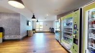 This single-floor communal space is thoughtfully designed to serve as a functional and inviting gathering area. The layout is divided into two primary zones: a lounge area and a convenience service area. The living room zone features a striking exposed concrete accent wall as its focal point, with a wall-mounted flat-screen television positioned above a flexible seating arrangement of colorful hexagonal ottomans and a low-profile coffee table. Wood-look plank flooring runs throughout the entire space, adding warmth and cohesion. Industrial-style pendant lights hang from the white ceiling, complementing the modern aesthetic. Large windows along one wall allow natural light to fill the interior while offering views of the surrounding outdoor greenery. Adjacent to the lounge, a dark-countered kitchenette station provides a small appliance area for additional convenience. The service zone houses a bank of canteen-style vending machines offering snacks and beverages, positioned near glass-paneled entry doors that connect to the exterior. The overall ambiance is contemporary and casual, balancing industrial design elements with approachable, functional furnishings suited for communal use.