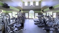 This fitness center is a spacious, single-floor facility designed to support a wide range of workout routines. The room immediately impresses with its soaring ceiling height, accentuated by a decorative arched window that serves as a striking focal point along the far wall. Large windows line the perimeter, flooding the interior with natural light and offering pleasant views of the surrounding greenery outside. A ceiling fan overhead provides additional air circulation throughout the space. The walls are finished in a bold two-tone color scheme of gray and green horizontal stripes, giving the facility a dynamic, energetic aesthetic. The flooring is a durable gray surface suited for high-traffic athletic use. On one side of the room, a comprehensive selection of strength training and cable machines is neatly arranged, catering to both upper and lower body workouts. The opposite side features a full row of cardio equipment, including treadmills, ellipticals, and stationary bikes. Wall-mounted televisions are positioned at multiple points for entertainment during workouts. The overall atmosphere is bright, open, and well-organized, making it a functional and motivating environment for fitness enthusiasts.