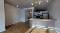 This modern single-floor unit showcases an industrial-inspired aesthetic that blends raw architectural elements with contemporary finishes. The open-concept living and kitchen area is immediately striking, defined by exposed concrete ceilings that lend an urban loft character to the space. Track lighting runs along the ceiling, providing flexible and functional illumination throughout.

The kitchen is positioned along one side of the open layout, featuring dark cabinetry, stainless steel appliances including a refrigerator, microwave, and gas range, as well as a built-in sink. A peninsula-style breakfast bar extends outward, offering additional workspace and casual seating while maintaining a seamless visual connection to the living area.

Warm hardwood flooring spans the entirety of the main living space, providing a natural contrast to the cool tones of the concrete ceiling and grey walls. The open floor plan allows for versatile furniture arrangements and creates a sense of spaciousness throughout. A hallway visible from the main living area leads to additional rooms beyond, suggesting a well-organized layout. The overall ambiance is clean, contemporary, and industrial — ideal for those who appreciate minimalist design with character-rich architectural details.
