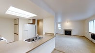 This single-floor residence offers a thoughtfully designed open-concept layout that seamlessly connects the kitchen and living areas. The kitchen is anchored by a peninsula counter with a double stainless steel sink, providing both workspace and a natural divider between the cooking and living zones. A skylight overhead floods the kitchen with natural light, complementing the white refrigerator and warm-toned upper cabinetry. The flooring transitions from the kitchen's hard surface to plush wall-to-wall carpeting in the living area, adding warmth and comfort underfoot. The living room is a welcoming space highlighted by a classic fireplace with a tile surround, serving as a natural focal point. A window allows additional natural light to filter into the living area, enhancing the open and airy atmosphere. Neutral white walls throughout keep the space feeling clean and versatile, ready to accommodate a variety of interior styles. A hallway visible from the kitchen suggests additional rooms beyond, completing the functional and comfortable layout of this well-appointed home.