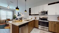 This single-floor residence showcases a thoughtfully designed open-concept layout with modern finishes throughout. The kitchen serves as a central highlight, featuring a two-tone cabinet design with warm wood lower cabinets and crisp white upper cabinets, complemented by open shelving. Granite countertops extend across the workspace and peninsula island, while a white subway tile backsplash adds a clean aesthetic. Stainless steel appliances, including a range, microwave, dishwasher, and refrigerator, complete the culinary space. Two pendant lights with warm-toned interiors hang above the island, adding both function and style. Light wood-style flooring runs throughout the main living areas, connecting the kitchen to the living room, where large windows allow natural light to fill the space. The bedroom offers carpeted flooring and generous window coverage with blinds for privacy. The bathroom presents a long vanity with granite countertops, warm wood cabinetry, and a tub-shower combination. Wood-look flooring ties the bathroom's design to the rest of the home, creating a cohesive and contemporary aesthetic throughout.