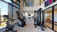 This contemporary fitness center is designed to deliver a premium workout experience within a thoughtfully crafted architectural environment. The main exercise floor impresses with soaring double-height ceilings and expansive floor-to-ceiling windows that flood the space with natural light and frame sweeping city skyline views. The neutral-toned carpet flooring and crisp white walls create a clean, focused atmosphere throughout.

The main workout area is comprehensively equipped with a strong lineup of Matrix-brand cardio machines, including stair climbers and additional aerobic equipment, alongside a robust selection of cable and strength training stations. Wall-mounted televisions are positioned throughout for easy viewing during workouts. Mirrored walls amplify the sense of space and support form-focused training.

A dedicated spin studio is enclosed by sleek floor-to-ceiling glass walls and a black-framed glass door, offering both visual openness and acoustic separation. Inside, a row of SoulCycle-style stationary bikes face panoramic windows overlooking the cityscape, making every cycling session visually engaging. A striking large-scale artistic mural in warm orange, teal, and white tones adorns the studio's exterior wall, adding a vibrant, creative focal point to the facility's corridor and reinforcing the space's modern, design-forward character.
