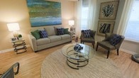 This single-floor space offers a welcoming and well-curated interior that balances comfort with contemporary style. The living area serves as the centerpiece of the home, drawing the eye immediately with its bold, large-scale abstract canvas in rich shades of blue, green, and gold, anchoring the room with a vibrant artistic focal point. Warm-toned walls complement the natural light streaming in through white-curtained windows, creating a bright and airy atmosphere.

The living room is furnished with a plush, neutral-toned sofa accented by an array of blue and green decorative pillows, adding layers of color and texture. A pair of sophisticated charcoal accent chairs with nail-head trim detail provides additional seating and a refined contrast to the lighter palette. At the center of the seating arrangement sits a striking round glass-top coffee table with a sculptural metal base, resting on a large circular woven area rug that grounds the space with organic texture.

Light hardwood flooring runs throughout, enhancing the overall warmth and openness of the interior. Framed botanical and landscape artwork adorns the walls, reinforcing the nature-inspired color story carried throughout the décor. The space is thoughtfully staged with carefully chosen accessories, reflecting a cohesive and inviting design aesthetic suited for both relaxed living and gracious entertaining.