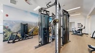 Located at 350 Artisan Dr in Laguna Beach, CA, this two-floor fitness facility offers a well-appointed and thoughtfully designed workout environment spread across two distinct levels. The upper floor houses a bright, open gym space with light wood flooring, recessed panel lighting, and ceiling fans. It is equipped with an extensive array of Matrix strength and cardio machines, including cable systems, treadmills, ellipticals, and spin bikes. Full-length mirrors amplify the sense of space, while large panoramic landscape murals depicting Southern California's iconic terrain lend a distinctive visual character. A dedicated free weights area features power racks, benches, and a motivational "ONE MORE" wall display. The lower level transitions into a high-intensity functional training zone with turf flooring and soaring ceiling height. This area includes a bouldering and rock climbing wall, sled push tracks, TRX suspension training stations, kettlebells, battle ropes, a punching bag, and a stair climber. Bold dark wall graphics reinforce the performance-focused atmosphere. Together, the two floors create a comprehensive facility that caters to a wide range of fitness disciplines, from traditional strength training to dynamic functional movement.