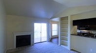 This single-floor residence centers around an open living area designed to maximize both functionality and natural light. The main living space features a carpeted floor and a fireplace framed in tile, providing a focal point along one wall. Vaulted ceilings add a sense of openness and volume to the room, enhancing its airy feel.

A sliding glass door opens directly to a private balcony with a metal railing, offering outdoor views and bringing abundant natural light into the space. An additional window in the adjacent area further brightens the interior, which transitions to hardwood or laminate flooring in the connected room.

Built-in shelving units flank a transitional wall near the main living area, offering generous display and storage space. The shelves are tall, reaching close to the ceiling, and are neatly integrated into the wall. Adjacent to the shelving, a glimpse of a countertop with dark stone or granite surfacing suggests a kitchen or dining area nearby.

The overall ambiance is light, open, and functional, with architectural details such as the vaulted ceiling and built-ins adding character to the otherwise neutral, move-in-ready interior.