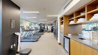 Located at 50 Minorca Ave in Coral Gables, Florida, this professionally designed fitness center offers a comprehensive exercise facility across a single, generously sized floor. The space is thoughtfully organized into distinct training zones, each outfitted with professional-grade equipment. A dedicated cardio area lines the perimeter, featuring multiple Matrix treadmills and elliptical trainers positioned alongside floor-to-ceiling windows that invite natural light and offer views of lush tropical palm trees outside. A strength training section provides a full rack of dumbbells and a multi-station cable machine, while a functional training area is stocked with kettlebells, medicine balls, stability balls, and TRX suspension trainers. Large wall-mounted mirrors throughout the space enhance the sense of openness and allow users to monitor their form. The flooring transitions between neutral-toned carpet and dark rubberized tiles, delineating each training zone clearly. Overhead, a combination of large circular pendant lights and recessed linear fixtures ensures bright, even illumination. A built-in display screen serves as a focal point in the functional zone. The clean, modern design features a crisp white ceiling, dark mosaic tile accent walls, and warm wood baseboards, creating a polished yet energizing atmosphere ideal for a complete workout experience.