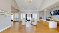 Located at 400 Claysburg Loop in Bloomingdale, Georgia, this thoughtfully designed single-floor space offers a welcoming and functional environment with a refined coastal aesthetic. The living room is a standout feature, boasting soaring vaulted ceilings with a shiplap finish that amplify the sense of openness. Large black-framed double-entry doors and expansive grid-pane windows flood the interior with natural light while providing views of the surrounding outdoor landscape. Warm-toned hardwood flooring flows seamlessly throughout, complementing the soft neutral wall palette. Comfortable seating arrangements throughout the living area invite gathering and relaxation, while a dedicated workspace nook — fitted with a built-in desk, white cabinetry, open wood shelving, and coastal-themed artwork — adds a practical yet stylish touch. The kitchen is equally impressive, featuring a generous center island with white shaker cabinetry, quartz countertops, a light blue subway tile backsplash, open wood shelving, stainless steel appliances, and gold pendant lighting. A wide window at the kitchen's end frames a bright outdoor view, further enhancing the airy ambiance. Together, these spaces create a cohesive, inviting environment ideal for community use.