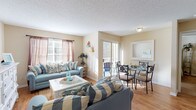 This single-floor unit showcases a warm, coastal-inspired aesthetic throughout its thoughtfully designed layout. The open-concept living and dining area serves as the heart of the home, where light hardwood flooring, warm beige walls, and abundant natural light from large windows create an inviting atmosphere. The living space features a seating arrangement with blue sofas accented by nautical-themed pillows, a white coffee table with a glass top, and a white dresser with louvered cabinet doors adding to the beachy charm.

The adjacent dining area accommodates a round glass-top table with seating for four and is positioned beside a sliding glass door that opens to a private outdoor balcony, allowing natural light to flow freely into the space. A decorative mirror on the wall adds depth to the dining nook.

The galley-style kitchen offers ample storage with warm wood cabinetry, dark countertops, a double-basin sink, and black appliances including a refrigerator and range. The bedroom is a restful retreat with plush carpeting, a ceiling fan with a light fixture, a louvered headboard, and a closet. The bathroom features wood cabinetry with dark countertops, a large vanity mirror, and a tiled shower-tub combination.