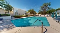 Located in Albuquerque, New Mexico, this outdoor recreational area showcases a generously sized swimming pool as its centerpiece, framed by a clean, sun-drenched concrete deck and surrounded by mature trees that provide natural shade and a sense of seclusion. The pool area is well-appointed with multiple rows of modern lounge chairs positioned along the deck, offering ample space for relaxation under the abundant New Mexico sunshine.

A standout feature of the space is a set of sleek, pergola-style cabana structures with slatted roofs, providing shaded retreats for loungers seeking relief from the sun. The surrounding landscape is thoughtfully maintained, with trimmed hedgerows, desert-friendly plantings, and gravel accents that reflect the regional aesthetic.

Access to the pool area is provided through a secured gate entrance framed by mature trees and ornamental fencing, ensuring privacy and safety. A dedicated building entry features decorative blue-and-white patterned tile risers on its steps, adding a distinctive Southwestern design touch. Nearby, a separate spa or hot tub area is accessible via similarly tiled stairways. The surrounding multi-story stucco architecture, consistent with the Pueblo Revival style common to Albuquerque, completes the cohesive and inviting ambiance of this well-maintained outdoor amenity space.