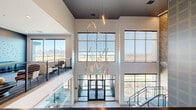 This two-story amenity facility in Bozeman, Montana makes a striking first impression with a soaring double-height lobby entrance, floor-to-ceiling glass walls, and a dramatic sculptural chandelier of cascading linear LED elements that spans both floors. Large windows frame sweeping views of the surrounding Montana landscape and distant mountain ranges, filling the space with abundant natural light.

The main level opens into a spacious lounge and game room outfitted with comfortable seating, a foosball table, and a communal bar-height island — ideal for casual gatherings. An adjacent kitchen area features dark navy cabinetry, a white tile backsplash, and direct access to an outdoor patio. A sleek staircase with glass-panel railings and textured accent walls connects the two levels.

The upper floor houses a dedicated business center offering a variety of work and meeting environments. A formal conference room features a substantial dark stone table, textured accent wallcovering, integrated AV, and frosted glass partitions for privacy. A library-inspired meeting room showcases a floor-to-ceiling built-in wood bookcase with warm gold accent lighting and a marble-topped table. An additional conference room offers a warm slatted wood ceiling, plaid upholstered seating, and a wall-mounted display — creating a refined yet inviting atmosphere throughout.