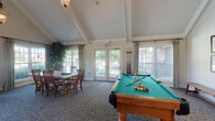 Located at 4311 Camden Cir in Dublin, Ohio, this single-story community clubhouse offers a thoughtfully designed gathering space that blends comfort with functionality. The interior is anchored by dramatic vaulted ceilings with exposed beam detailing and elegant pendant lighting, creating an open and airy atmosphere throughout.

The main lounge area is a standout feature, centered around a stately stone fireplace with a mounted television above. Plush neutral sofas and accent chairs are arranged to encourage relaxed socializing, while large windows dressed in floor-length drapes invite abundant natural light and views of the surrounding grounds.

Adjacent to the lounge, a reception counter provides a dedicated service area, seamlessly integrated into the open floor plan. The billiards room is a highlight for recreation, housing a full-size pool table beneath a soaring vaulted ceiling section, accompanied by a casual dining table and chairs for additional gathering space.

A dedicated dining area features a large rustic wood table surrounded by classic wooden chairs, positioned near French doors that open directly to the outdoors, enhancing the connection between interior and exterior spaces. Multiple windows throughout ensure the entire clubhouse remains bright and welcoming. Patterned carpeting ties the spaces together, contributing to the warm and inviting character of this community amenity.