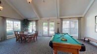Located at 4311 Camden Cir in Dublin, Ohio, this single-story community clubhouse offers a thoughtfully designed gathering space that blends comfort with functionality. The interior is anchored by dramatic vaulted ceilings with exposed beam detailing and elegant pendant lighting, creating an open and airy atmosphere throughout.

The main lounge area is a standout feature, centered around a stately stone fireplace with a mounted television above. Plush neutral sofas and accent chairs are arranged to encourage relaxed socializing, while large windows dressed in floor-length drapes invite abundant natural light and views of the surrounding grounds.

Adjacent to the lounge, a reception counter provides a dedicated service area, seamlessly integrated into the open floor plan. The billiards room is a highlight for recreation, housing a full-size pool table beneath a soaring vaulted ceiling section, accompanied by a casual dining table and chairs for additional gathering space.

A dedicated dining area features a large rustic wood table surrounded by classic wooden chairs, positioned near French doors that open directly to the outdoors, enhancing the connection between interior and exterior spaces. Multiple windows throughout ensure the entire clubhouse remains bright and welcoming. Patterned carpeting ties the spaces together, contributing to the warm and inviting character of this community amenity.