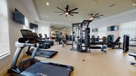 This fitness center is a thoroughly equipped exercise facility designed to support a wide variety of workout routines. The space features a light-toned, durable rubberized floor and crisp white walls accented by recessed lighting and large ceiling fans that maintain airflow throughout the room. Natural light flows in generously through multiple windows fitted with white blinds, creating a bright and energizing atmosphere. A full-length wall mirror on one end visually expands the space and allows users to monitor their form during workouts. The cardio section is stocked with True-brand treadmills, rowing machines, and ellipticals, while the strength training area features an Atlas cable machine system, a TKO heavy punching bag, multiple selectorized weight machines for targeted muscle groups, and adjustable benches. An Endurance-branded piece of equipment adds further versatility. A dedicated functional training zone includes stability balls on a stand, additional benches, and open floor space for stretching or bodyweight exercises. Wall-mounted flat-screen televisions are positioned throughout the room for entertainment during workouts. Built-in cabinetry along one wall provides storage. The overall design is clean, modern, and purposefully organized to accommodate a full-spectrum fitness experience.