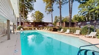 Located at 50 Chateau Dr SE in Rome, Georgia, this outdoor pool area offers a well-appointed recreational retreat set against a lush natural landscape. The centerpiece is a generously sized rectangular swimming pool with clear blue water, accessible via handrailed entry steps and surrounded by a wide concrete pool deck. The deck is furnished with multiple white lounge chairs and patio seating arrangements, complemented by purple patio umbrellas that provide shaded areas for relaxation on sunny days. A covered area along one side of the pool features attractive stone masonry accents on the adjacent building exterior, offering a sheltered space near the water's edge. The surrounding grounds are landscaped with neatly trimmed hedges and mature, towering pine trees that frame the space and create a serene, park-like ambiance. A fenced perimeter encloses the pool area, providing privacy and security. Residential buildings with wood-sided exteriors are visible in the background, integrating the pool area naturally into the broader community setting. The space is bathed in abundant natural light, making it an inviting outdoor amenity well-suited for leisure and recreation throughout the warmer months.