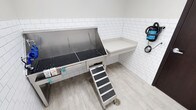 This purpose-built pet grooming space is designed with both functionality and cleanliness in mind. The room is finished with classic white subway tile on the walls and smooth, wood-look flooring, creating a bright and hygienic environment. At the center of the space sits a professional-grade stainless steel grooming tub equipped with a non-slip rubber grate base, integrated storage drawers, and grooming supply organizers — ideal for accommodating pets of various sizes. A set of anti-slip entry steps provides safe and easy access to the tub, making the experience comfortable for animals. Adjacent to the tub, a wall-mounted white work shelf offers a convenient flat surface for grooming tools and supplies. A wall-mounted pet dryer with a flexible hose keeps everything within easy reach without cluttering the workspace. A floor drain is positioned nearby for easy cleanup and maintenance. The overall layout is thoughtfully organized to maximize efficiency, with every element oriented toward delivering a professional grooming experience. The neutral color palette, clean lines, and dedicated fixtures make this a well-equipped and welcoming space for pet care.