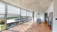 This contemporary high-rise unit showcases an industrial-modern aesthetic defined by soaring exposed concrete ceilings and expansive floor-to-ceiling windows that flood the interior with natural light. The open-concept living and dining area is anchored by warm hardwood flooring and a dramatic wall of glass offering sweeping panoramic views of the surrounding cityscape and treetops. Structural concrete columns add an architectural character that complements the unit's modern design language.

The kitchen is thoughtfully appointed with two-tone cabinetry — crisp white upper cabinets paired with gray lower cabinets — white subway tile backsplash, quartz countertops, and a full suite of stainless steel appliances. A spacious center island with seating provides additional prep and dining space, while sleek pendant lighting adds a stylish finishing touch overhead.

The bedrooms are carpeted for comfort and feature large windows with roller shades that continue the unit's bright, airy feel. The bathrooms are finished with wood-look luxury vinyl flooring, gray vanity cabinetry, and clean white fixtures, with one bathroom featuring accessibility grab bars. A dedicated in-unit laundry closet houses a stacked washer and dryer with built-in shelving above for added storage convenience.