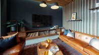 This single-floor communal lounge is a thoughtfully designed space that blends mid-century modern aesthetics with contemporary finishes. Slatted wood paneling runs across both walls and ceilings throughout, creating a warm, rhythmic texture that unifies the open layout. Polished concrete floors provide an industrial counterpoint, grounding the space with understated sophistication.

The lounge area anchors one end of the space, featuring rich cognac leather sofas atop vibrant vintage-style area rugs. A large flat-screen television is mounted above a custom walnut media console housing an extensive vinyl record collection, making it a clear focal point for music and entertainment enthusiasts. Floating shelves display curated artwork, framed prints, and greenery, adding personality and visual interest.

A full-length ping-pong table takes center stage in the main open area, positioned beneath a slatted wood ceiling fitted with recessed and pendant lighting. Adjacent to it, a stylish wet bar finished in deep teal zellige tile serves as a social hub, complete with open shelving and a wood-topped counter.

Floor-to-ceiling windows and glass sliding doors flood the space with natural light and open to an exterior balcony, offering views of the surrounding urban environment. Curated artwork punctuates the walls throughout, reinforcing the space's creative and social character.