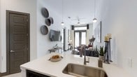This single-story, one-bedroom, one-bathroom unit features a thoughtfully designed open-concept floor plan that maximizes the available living space. The kitchen flows seamlessly into the living area, anchored by a sleek countertop with an undermount stainless steel sink and a contemporary gooseneck faucet. Light-toned quartz or stone counters provide ample prep space, while pendant globe lights hang above, adding a modern aesthetic touch.

The living area extends toward the back of the unit, where large floor-to-ceiling glass doors allow abundant natural light to filter in, creating an airy and open atmosphere. A ceiling fan with integrated lighting serves the main living space, enhancing both comfort and functionality. The walls are painted in a clean, neutral white tone, providing a versatile backdrop for décor.

Wall-mounted television placement and built-in shelving options offer practical media solutions without compromising the sense of space. The unit's linear layout guides the eye naturally from the kitchen through to the living area, making the space feel expansive. Overall, the unit presents a clean, contemporary aesthetic well-suited for modern urban living.