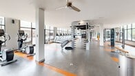 Located at 3106 Jacobs Rd in Seneca, SC, this single-floor fitness center offers a generously sized, well-equipped workout facility designed to support a comprehensive range of exercise disciplines. The open floor plan is finished with durable, dual-tone rubber flooring in grey and orange, providing both visual energy and practical resilience underfoot.

The cardio zone features a row of treadmills and elliptical machines positioned along expansive floor-to-ceiling windows, allowing natural light to flood the space and providing clear views of the surrounding campus-style grounds. Adjacent to the cardio area, a dedicated spin studio houses multiple Spinning-brand stationary bikes and is equipped with a wall-mounted display screen, creating an immersive cycling environment.

The strength training area is anchored by a large multi-functional cable and suspension training rig, complemented by a full rack of dumbbells, kettlebells, medicine balls, and various free weight benches. Structural columns are integrated thoughtfully throughout, supporting ceiling fans that enhance air circulation across the expansive floor area.

The facility maintains a clean, modern aesthetic with bright white walls, recessed and flush-mounted lighting, and multiple access points including an exterior door. The overall ambiance is functional and motivating, suited for both individual training and group fitness activities.