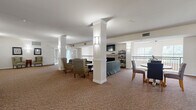 Located at 1745 Graham Ave in Saint Paul, MN, this generously proportioned greatroom community space spans a single floor and offers a thoughtfully designed layout for socializing and recreation. The main lounge area is anchored by prominent white columns that define the space while maintaining an open, airy feel. Comfortable seating arrangements — including striped armchairs and a plush sofa — are positioned around a fireplace with a mounted television, creating an inviting gathering spot. Wall sconces provide warm ambient lighting throughout, complementing the neutral patterned carpeting that flows across the entire floor. Large windows allow natural light to fill the space and offer views of the surrounding exterior. An elegant staircase with ornate dark metal railings and a chandelier connects to adjacent areas, adding a refined architectural touch. The hallway extends the usable space and features additional seating nooks bathed in natural light. The dedicated game room is a standout feature, equipped with a full-size billiard table set against cool blue accent walls with skylights overhead, as well as a ping-pong table in a separate open area flanked by white columns, track lighting, and greenery. The overall ambiance is welcoming, functional, and well-appointed.