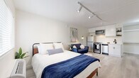 This thoughtfully designed studio offers a seamless open-plan layout that efficiently combines the sleeping, living, and dining areas into a single cohesive space. Light-toned hardwood flooring runs throughout, lending warmth and a sense of continuity, while crisp white walls and track lighting overhead create a bright, clean aesthetic.

The sleeping and living area forms the heart of the studio, comfortably accommodating a full-size bed and a compact seating area. A wall-mounted flat-screen television adds a modern entertainment feature, and a natural light source via a window keeps the space feeling airy and open. A bold teal entry door adds a striking design accent upon entry.

The kitchen, positioned along one wall, is fully equipped with stainless steel appliances including a range, microwave, refrigerator, and dishwasher. White shaker-style cabinetry paired with white quartz countertops and a matte black faucet give the kitchen a clean, contemporary look with practical functionality.

The bathroom is finished to a high standard, featuring large-format slate-grey floor tiles, a glass-enclosed walk-in shower with a rain showerhead, a floating vanity with a white countertop, and matte black fixtures throughout — delivering a spa-like atmosphere in a compact footprint. Overall, the studio presents a modern, efficient, and well-finished living environment.