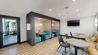 Located at 575 Matmor Rd in Woodland, CA, this thoughtfully designed single-floor communal space offers a cohesive blend of modern aesthetics and functional layout. The kitchen is a standout feature, showcasing bold blue cabinetry, open floating wood shelves with integrated lighting, a subway tile backsplash, and a built-in oven. A wood-topped bar-height island with black modern stools serves as both a prep and casual dining area, illuminated by stylish gold and white pendant lights suspended from a rustic exposed wood beam. Natural light pours in through a large picture window overlooking lush outdoor greenery. The lounge and seating areas extend across light wood vinyl plank flooring, featuring a mix of a tan leather banquette with round tables, semi-private teal booth seating in a dedicated nook, and additional open seating around circular tables. A wall-mounted flat-screen TV and decorative abstract artwork with warm sconce lighting add to the polished, inviting atmosphere. Black-framed glass doors connect the space to adjacent areas, enhancing the open, accessible feel of this well-appointed communal environment.