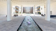 This expansive outdoor courtyard amenity space is the centerpiece of a modern residential complex, framed on multiple sides by multi-story buildings with balconied facades in brick and light stone. A long, linear water feature runs through the heart of the courtyard, with illuminated fountains creating an eye-catching focal point that draws the eye toward a full-size resort-style swimming pool at the far end. The pool deck is lined with lounge chairs, offering a dedicated space for relaxation under open skies. Overhead, strings of café lights are strung across the courtyard, lending a warm, inviting ambiance during evening hours. A covered pavilion area anchors one end of the space, supported by tall white columns and featuring a soaring ceiling equipped with a statement ceiling fan. The pavilion opens onto the main courtyard through large glass doors, connecting the interior amenity spaces to the outdoors seamlessly. The entire ground surface is finished with a carefully laid paver pattern, adding a refined, urban aesthetic throughout. Outdoor seating arrangements, including lounge chairs and dining table sets, are thoughtfully distributed across the space, accommodating both casual gatherings and more formal outdoor dining. The overall design strikes a balance between modern architectural elegance and functional outdoor living.