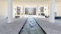 This expansive outdoor courtyard amenity space is the centerpiece of a modern residential complex, framed on multiple sides by multi-story buildings with balconied facades in brick and light stone. A long, linear water feature runs through the heart of the courtyard, with illuminated fountains creating an eye-catching focal point that draws the eye toward a full-size resort-style swimming pool at the far end. The pool deck is lined with lounge chairs, offering a dedicated space for relaxation under open skies. Overhead, strings of café lights are strung across the courtyard, lending a warm, inviting ambiance during evening hours. A covered pavilion area anchors one end of the space, supported by tall white columns and featuring a soaring ceiling equipped with a statement ceiling fan. The pavilion opens onto the main courtyard through large glass doors, connecting the interior amenity spaces to the outdoors seamlessly. The entire ground surface is finished with a carefully laid paver pattern, adding a refined, urban aesthetic throughout. Outdoor seating arrangements, including lounge chairs and dining table sets, are thoughtfully distributed across the space, accommodating both casual gatherings and more formal outdoor dining. The overall design strikes a balance between modern architectural elegance and functional outdoor living.