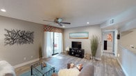 This single-floor residence offers a welcoming and well-appointed living space designed with both comfort and style in mind. The living room serves as the heart of the home, featuring wood-look laminate flooring that runs throughout and adds warmth and continuity to the space. A textured ceiling with a centrally mounted ceiling fan provides both character and airflow, while recessed lighting ensures the room is well-lit at all times.

Natural light pours in through large sliding glass doors dressed with sheer white curtains and a vibrant geometric-patterned valance, creating a bright and airy atmosphere. The doors open to an outdoor area, extending the living space beyond the interior. Neutral gray walls serve as a versatile backdrop, complementing the contemporary furnishings that include a glass-top coffee table and a plush sofa accented with decorative pillows.

Tall decorative grasses and wall art add organic and coastal-inspired touches, enhancing the relaxed ambiance of the space. A media console with a flat-screen television anchors one wall, making the living room equally suited for entertaining or everyday relaxation. A hallway visible from the main area suggests additional rooms, contributing to the overall functional layout of this thoughtfully designed residence.