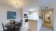This single-floor residence showcases a thoughtfully arranged open-concept layout connecting the kitchen and dining areas seamlessly. The kitchen is equipped with essential appliances, including a white refrigerator, electric range, and built-in microwave, all framed by white cabinetry that contributes to a clean, bright aesthetic. A peninsula counter extends into the living space, offering additional prep area while reinforcing the open flow between rooms. Recessed flush-mount ceiling lighting illuminates the kitchen efficiently. The adjacent dining area features a classic chandelier with candelabra-style lights, adding a touch of traditional elegance. A dark wood dining table with matching chairs anchors the space, complemented by decorative wall art and potted plants that introduce natural color. Neutral beige walls and carpeted flooring carry throughout, creating a warm and cohesive atmosphere. A hallway visible from the main space suggests additional rooms beyond, contributing to the overall functional layout of this well-organized single-floor home.