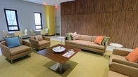 This single-floor space presents a well-appointed lounge or common area designed with a clean, contemporary aesthetic. The room's most striking architectural feature is a floor-to-ceiling wood-paneled accent wall, which adds warmth and visual depth to the space while serving as a bold focal point. The seating arrangement is thoughtfully composed, centered around a large, neutral-toned sofa accented with colorful throw pillows in shades of orange, green, and teal, complemented by matching armchairs that frame the space comfortably. A low-profile wooden coffee table with a metallic base anchors the seating group, topped with decorative accents and reading materials. The flooring features a muted green carpet that ties the color palette together cohesively. Adjacent to the main lounge area, a casual dining or breakout nook is visible, furnished with light-toned chairs and a small table near a window that brings in natural light. The yellow-painted walls in that alcove introduce a cheerful contrast to the wood and neutral tones of the main area, contributing to an overall atmosphere that is modern, inviting, and functional.