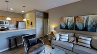 This single-floor space showcases a thoughtfully designed open-concept layout that seamlessly connects the living area to the kitchen. The living room is anchored by a plush gray sofa accented with decorative pillows and paired with a dark upholstered armchair, creating a comfortable and stylish seating arrangement. A soft area rug defines the space underfoot, while dark hardwood-style flooring extends throughout, adding warmth and visual continuity. A large abstract canvas painting serves as a focal point on the taupe-toned accent wall, complementing the neutral color palette of warm grays and beiges used throughout. Ambient lighting from wall-mounted fixtures and a small accent lamp contributes to the cozy atmosphere. The kitchen is accessible via a raised breakfast bar pass-through, with pendant lighting overhead and light-colored countertops visible from the living area. White appliances and warm wood cabinetry complete the kitchen's classic aesthetic. A white entry door with adjacent wall shelf provides a functional entryway transition. The space is compact yet efficiently arranged, making excellent use of every area to balance comfort, style, and practicality.