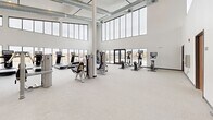 Located at 303 Airway Blvd in Belgrade, Montana, this well-appointed fitness center offers a generously sized, single-floor exercise space designed to support a comprehensive workout experience. The facility's most striking architectural feature is its dramatic double-height ceiling, which creates an open, airy atmosphere throughout the entire room. Exposed industrial ductwork runs overhead, complementing the building's modern aesthetic while adding a clean, functional character.

Floor-to-ceiling windows line multiple walls, flooding the interior with abundant natural light and framing sweeping views of the surrounding Montana landscape, including snow-capped mountain ranges and an adjacent outdoor pool and patio area. The light-toned carpet flooring and crisp white walls further enhance the bright, welcoming environment.

The exercise room is divided into distinct functional zones. The cardio area is equipped with treadmills, stationary bikes, and elliptical machines, many featuring integrated display screens. A dedicated strength training section includes a full rack of dumbbells, adjustable benches, and a variety of cable and resistance machines. Full-length mirrors along one wall visually expand the space and provide practical feedback during workouts.

Large glass doors provide direct access to the outdoor pool area, seamlessly connecting indoor and outdoor amenities. The overall design is clean, contemporary, and purpose-built to deliver a premium fitness experience.