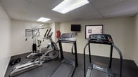 This self-contained fitness room offers a practical and well-equipped space for residents or guests to maintain their exercise routines. The room features a drop ceiling with recessed fluorescent panel lighting that provides bright, even illumination throughout. The flooring consists of durable, dark carpet with rubber mat sections placed beneath the equipment for added stability and protection.

The cardio section includes multiple treadmills — among them a PaceMaster and a Landice model — as well as an elliptical cross-trainer, offering a solid range of aerobic workout options. A wall-mounted flat-screen television is positioned to face the cardio equipment, allowing users to watch content during their sessions. A large wall mirror spans one side of the room, visually expanding the space and allowing users to monitor their form.

For strength training, the room features a multi-station cable weight machine complete with a weight stack, pull-down bar, and various attachment points suitable for a range of resistance exercises. A wall-mounted air conditioning unit provides climate control, ensuring comfort during workouts. Safety equipment, including a fire extinguisher, is also present. A window allows a modest amount of natural light to supplement the overhead lighting. The room is accessed through a dedicated entrance door, making it a clearly defined, purpose-built fitness facility.
