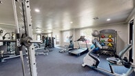 This fitness center at 5808 E 71st St in Tulsa, Oklahoma, offers a thoughtfully organized exercise space designed to support a full range of workout routines. The room features durable dark-toned carpet flooring throughout and is illuminated by a combination of recessed ceiling lights and natural light streaming through multiple windows, creating a bright and energizing atmosphere.

The cardio section is well-stocked with a Spirit treadmill, a Precor elliptical, and a stationary upright bike, providing varied options for aerobic training. For strength training, the space includes a multi-functional cable machine, a weight bench, a full rack of dumbbells in graduated sizes, and a TRX suspension training system. Additional fitness accessories such as medicine balls, stability balls, and a ball rack are neatly arranged along the walls.

A large wall-mounted mirror spans one side of the room, visually expanding the space and allowing users to monitor their form during workouts. A wall-mounted flat-screen television adds convenience for entertainment during exercise sessions. The space also features a glass door and sliding glass panels that connect to an adjacent studio area, offering flexibility for group fitness or additional training activities. Natural light from surrounding windows provides views of the outdoor environment, contributing to an open and motivating workout setting.
