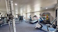 This fitness center at 5808 E 71st St in Tulsa, Oklahoma, offers a thoughtfully organized exercise space designed to support a full range of workout routines. The room features durable dark-toned carpet flooring throughout and is illuminated by a combination of recessed ceiling lights and natural light streaming through multiple windows, creating a bright and energizing atmosphere.

The cardio section is well-stocked with a Spirit treadmill, a Precor elliptical, and a stationary upright bike, providing varied options for aerobic training. For strength training, the space includes a multi-functional cable machine, a weight bench, a full rack of dumbbells in graduated sizes, and a TRX suspension training system. Additional fitness accessories such as medicine balls, stability balls, and a ball rack are neatly arranged along the walls.

A large wall-mounted mirror spans one side of the room, visually expanding the space and allowing users to monitor their form during workouts. A wall-mounted flat-screen television adds convenience for entertainment during exercise sessions. The space also features a glass door and sliding glass panels that connect to an adjacent studio area, offering flexibility for group fitness or additional training activities. Natural light from surrounding windows provides views of the outdoor environment, contributing to an open and motivating workout setting.