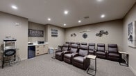 This private home theater and lounge is purpose-built for an immersive cinematic experience. The room is thoughtfully designed with two rows of plush, dark brown leather recliner chairs arranged to face a large wall-mounted flat-screen television paired with a soundbar, ensuring excellent sightlines from every seat. Small side tables positioned between the recliners provide convenient surfaces for snacks and drinks during a show.

The space is finished with neutral warm-taupe walls and soft carpeted flooring, creating a comfortable and acoustically friendly environment. Recessed ceiling lighting illuminates the room evenly, while the overall layout maximizes the sense of a true theater setting.

A dedicated snack and refreshment station lines one wall, featuring light wood cabinetry, a microwave, and a beverage refrigerator — everything needed for a complete at-home movie night. Cinema-themed wall décor adds personality throughout, including film reel accents, a "Lights Camera Action" sign, classic movie artwork, and a "Theater & Lounge" marquee sign that sets the tone from the moment you enter.

The room is accessed through a single door and offers an intimate, enclosed atmosphere ideal for screenings, gaming, or casual lounging. Every detail of this space has been curated to deliver a genuine theater experience in a private, comfortable setting.