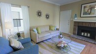 This single-story space showcases a thoughtfully designed living room that serves as its central feature. The room is dressed in warm, soft yellow walls that create an inviting and airy atmosphere, complemented by crisp white crown molding that adds a touch of classic architectural detail. Natural light filters in through a window dressed with sheer white curtains, brightening the space and enhancing its open feel.

The living room is anchored by a generously sized neutral-toned sofa adorned with green and blue accent pillows, paired with a coordinating blue upholstered chair. A round glass coffee table with a metal frame sits atop a striped area rug in muted green and grey tones, adding visual texture to the hardwood flooring beneath. Two decorative starburst mirrors mounted on the feature wall serve as eye-catching focal points.

A white interior door is visible toward the rear of the room, alongside a fireplace with a white mantel, adding a charming architectural element. The overall ambiance is light, casual, and well-balanced, making this a comfortable and functional living space.