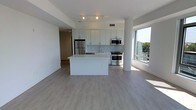 This single-floor unit showcases a clean, contemporary design with an open-concept layout that seamlessly connects the kitchen and living areas. The kitchen is outfitted with white flat-panel cabinetry, a subway tile backsplash, and a full suite of stainless steel appliances, including a refrigerator, gas range, and microwave. A central white island provides additional counter space and serves as a natural focal point, ideal for both meal preparation and casual dining.

Light gray luxury vinyl plank flooring runs throughout, lending a cohesive, modern aesthetic to the space. Track lighting on the ceiling ensures the interior is well-lit, complementing the abundant natural light that pours in through the large floor-to-ceiling windows along one wall. These windows also offer views of the surrounding greenery and suggest an elevated position within the building.

A cylindrical column near the window adds an architectural detail, while an open corridor leads toward additional rooms, hinting at further living space beyond the main area. The overall ambiance is bright, airy, and minimalist.