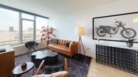 This thoughtfully designed single-floor residence offers a cohesive blend of modern aesthetics and functional living. The open-concept living room is a standout space, anchored by floor-to-ceiling windows that frame expansive city and urban waterfront views, flooding the room with natural light. Light hardwood flooring runs throughout, complementing the clean white walls and contemporary furnishings. A tan leather sofa, geometric coffee table, and a wooden media console with a wall-mounted television give the living area a stylish yet comfortable character.

The kitchen is equipped with stainless steel appliances, sleek white cabinetry, and a large central island topped with a gray quartz-style countertop, accommodating bar seating for two — ideal for casual dining or entertaining. Pendant lighting above the island adds a refined touch.

The bedroom features a generous walk-in closet and large windows that continue the theme of abundant natural light and urban views. The bathroom is finished with clean, modern materials, including large-format floor tiles, a frameless glass-enclosed walk-in shower lined with crisp white subway tiles, and a floating gray vanity with a white countertop. Together, these spaces create a well-proportioned, contemporary residence with a polished, urban sensibility.