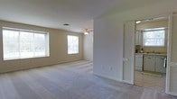 This single-floor residence offers a straightforward and functional layout designed for comfortable everyday living. The open living area is generously proportioned, featuring neutral-toned carpet flooring and warm beige walls that create a calm, understated ambiance throughout. Two windows fitted with white horizontal blinds allow ample natural light to fill the space, enhancing its airy and welcoming feel. A ceiling fan is centrally positioned overhead, providing both light and air circulation to the main living area. The open-concept flow connects the living space seamlessly to the kitchen area, which is visible from the main room and features white cabinetry, including louvered cabinet doors that add a subtle architectural detail. The kitchen is neatly appointed with white appliances and offers a practical workspace. A door adjacent to the kitchen provides additional access or storage options. The overall color palette is light and consistent — white trim, beige walls, and soft carpet — lending the space a cohesive and tidy appearance. The layout is efficient and well-lit, making it a practical and move-in ready living environment suited to a range of occupants.