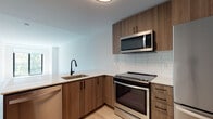 This single-floor unit showcases a thoughtfully designed open layout with a clean, modern aesthetic throughout. The kitchen serves as a functional centerpiece, featuring flat-front wood-grain cabinetry with matte black hardware, white quartz countertops, and a white subway tile backsplash. A full suite of stainless steel Whirlpool appliances includes a French door refrigerator, slide-in electric range, over-the-range microwave, and dishwasher. The kitchen opens to the living area via a peninsula with an integrated sink, creating a seamless flow between cooking and living spaces. Light wood-look flooring extends throughout the unit, enhancing the bright, airy atmosphere. The living area benefits from a large window that invites natural light and provides outdoor views. The bathroom features large-format light tile flooring, a wood-grain vanity with a neutral countertop, a wide illuminated mirror, and a clean white toilet. A generously sized walk-in closet equipped with wire shelving and hanging rods provides practical storage. The entry door features a warm wood finish, adding a welcoming touch to the overall contemporary design.