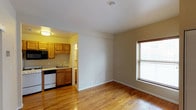 Located at 1030 N College in Indianapolis, Indiana, this single-floor space offers a clean, functional layout designed for comfortable everyday living. The main living area is open and bright, with warm-toned hardwood floors that run throughout and a large window fitted with horizontal blinds that allows natural light to fill the room, creating an inviting and airy atmosphere.

Adjacent to the living area, the kitchen is efficiently designed and features warm honey-toned wood cabinetry that provides ample storage space. The kitchen is equipped with a black built-in microwave, a dishwasher, and a sink, all set against light countertops. A doorway from the kitchen leads further into the unit, suggesting additional utility or storage space beyond.

The overall color palette is neutral and understated, with soft beige walls and white ceilings that complement the natural wood tones of the flooring and cabinetry. The space maintains a cohesive, practical aesthetic throughout. Clean lines and a straightforward layout make this unit easy to navigate and versatile for a variety of furnishing arrangements, making it a well-appointed option in the Indianapolis area.