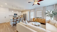 This two-story home is designed with a clean, contemporary aesthetic throughout, characterized by a neutral palette of whites and warm tones complemented by light hardwood-style flooring. The main level centers around an open-concept living and kitchen area that flows seamlessly together. The kitchen is outfitted with crisp white cabinetry, stainless steel appliances including a range and microwave, and a large central island with a white quartz countertop and seating for multiple bar-height chairs. Three pendant lights with glass shades hang above the island, adding a refined touch. Recessed lighting and natural light from nearby windows keep the space bright and welcoming.

The living room offers generous space for seating and features a ceiling fan, large windows overlooking the exterior, and access to a staircase leading to the upper level. The primary bedroom upstairs is distinguished by vaulted ceilings, dual windows, a ceiling fan, and a spacious walk-in closet with wire shelving systems. The en-suite bathroom features a wide double-sink vanity with quartz countertops, white cabinetry, and a glass-enclosed shower. A secondary bedroom offers natural light and ample closet space, while a full bath with a tub-shower combo serves additional occupants. A dedicated laundry room with full-size washer and dryer completes the home.