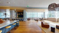 This expansive single-floor space is designed with a sophisticated, modern aesthetic that seamlessly blends open social areas with distinct functional zones. Warm hardwood flooring flows throughout, unifying the various areas while providing a natural contrast to the contemporary finishes.

A dramatic corridor lined with floor-to-ceiling glass partitions and sheer drapes leads into the main areas, where soaring ceilings amplify the sense of volume. The primary lounge features an extensive seating arrangement bathed in natural light from large-format windows overlooking a city skyline and lush palm trees. A live-edge communal dining table anchors one end, beneath a wood-paneled ceiling with recessed lighting.

A dedicated games room houses a full-size billiards table beneath a statement woven pendant light, accompanied by built-in banquette seating and a nearby wet bar with bar stool seating, wine storage, and open shelving. A striking putting green room features floor-to-ceiling moss-covered walls, offering a unique recreational retreat.

Throughout the space, curated artwork and sculptural furnishings add cultural depth, while a built-in bar with counter seating, open shelving, and a live green wall installation contribute to the vibrant, resort-style ambiance.