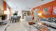 This stylish single-floor residence showcases a thoughtfully designed open-plan layout that connects the living, dining, and kitchen areas into one cohesive and inviting space. The interior is immediately striking, anchored by a bold terracotta-orange accent wall that adds warmth and personality to the open living area, contrasting beautifully with the crisp white ceilings and light-toned large-format floor tiles that run throughout.

The modern kitchen is equipped with stainless steel appliances, including a refrigerator and microwave, and features sleek taupe cabinetry with ample storage. A white kitchen island with bar-height seating serves as both a functional workspace and a casual dining spot, making it ideal for entertaining.

Adjacent to the kitchen, the dining area is defined by a distinctive round table paired with bold black-and-white striped chairs and a decorative wall mirror, lending an eclectic, artistic touch. The living area is furnished with a plush sofa, glass coffee table, and a soft area rug, creating a comfortable and visually dynamic gathering space.

Natural light fills the interior, enhanced by recessed lighting and stylish lamps throughout. The overall ambiance is modern and energetic, blending vibrant color choices with contemporary furnishings for a space that feels both curated and welcoming.