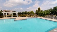 This outdoor amenity space centers around a generously sized swimming pool with clear, vibrant turquoise water, designed to accommodate both leisure and recreation. The pool deck is finished with clean, light-colored concrete surround, providing ample space for lounging and movement around the water's edge. A shallow wading area with step entry is conveniently situated adjacent to the main pool, offering easy water access for all users.

A standout architectural feature is the elegant white pergola positioned at the far end of the pool area. It serves as a shaded gathering spot and is flanked by outdoor seating and tables, creating an inviting space to relax and socialize. Potted flowering plants are strategically placed around the deck, adding vibrant splashes of color and a welcoming, well-maintained aesthetic.

The entire pool area is enclosed by a black iron fence, which provides both security and an unobstructed view of the picturesque pond situated just beyond the perimeter. Mature trees line the background, offering natural shade and a lush green backdrop. Classic lamp posts punctuate the space, suggesting the area is also functional during evening hours. The overall ambiance is that of a polished, resort-style community amenity designed for relaxation and outdoor enjoyment.